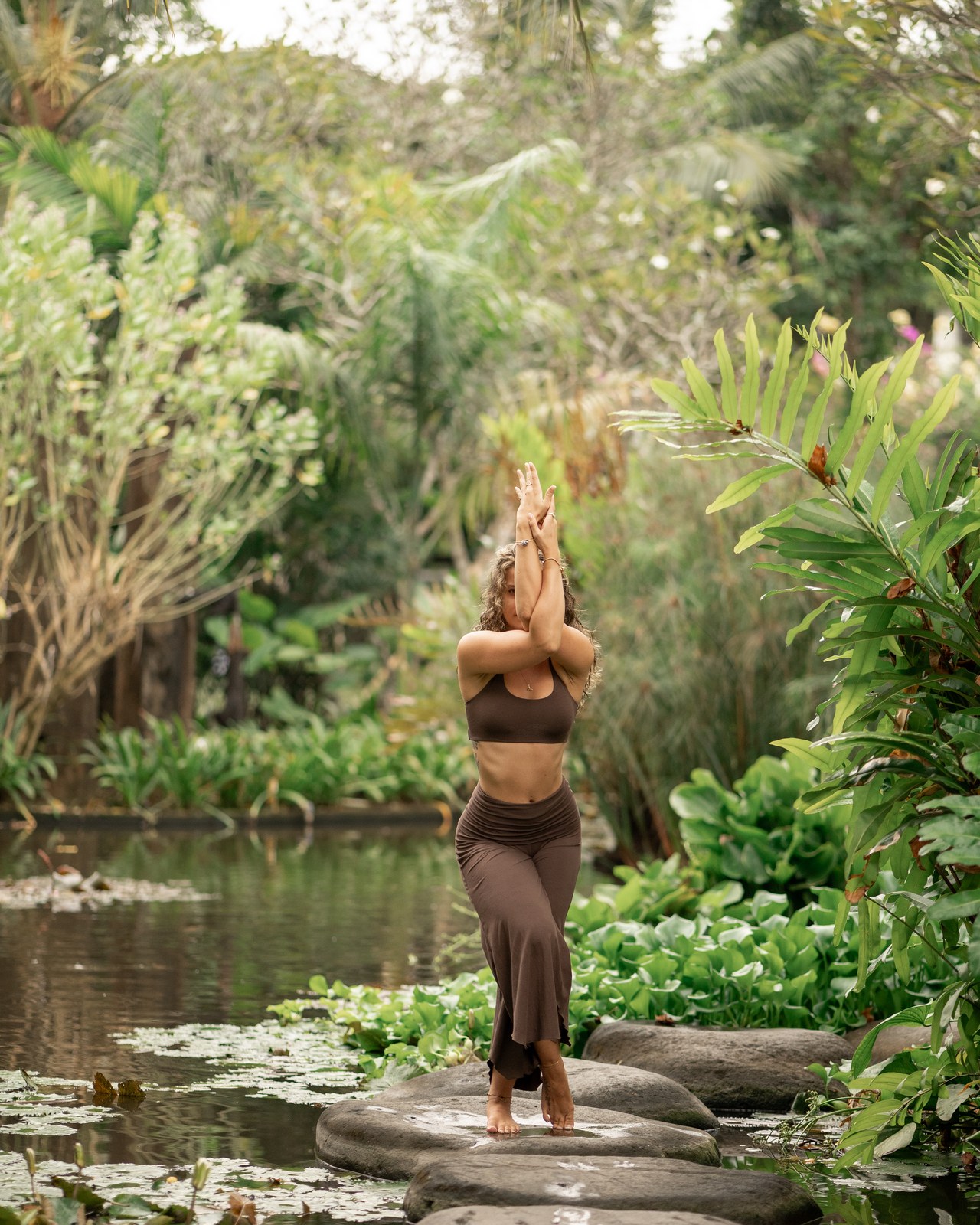 Josee standing with arms raised on stepping stones beside a tropical pond